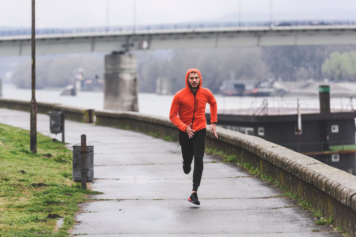 Handsome young athlete man running fast along river in orange windbreaker jacket.