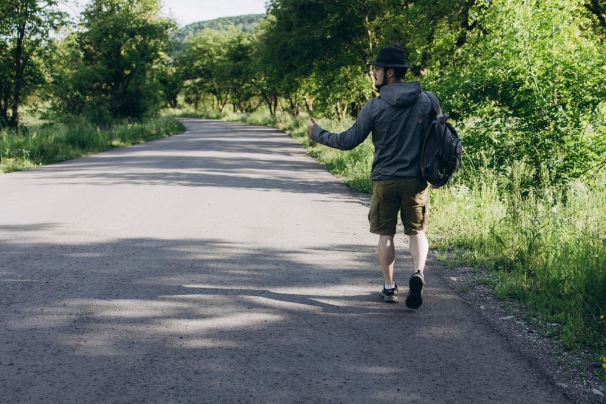 A man in shorts, a gray jacket and a hat hitchhikes car in road. Time to travel and auto stop concept. Green grass and sky after rain.