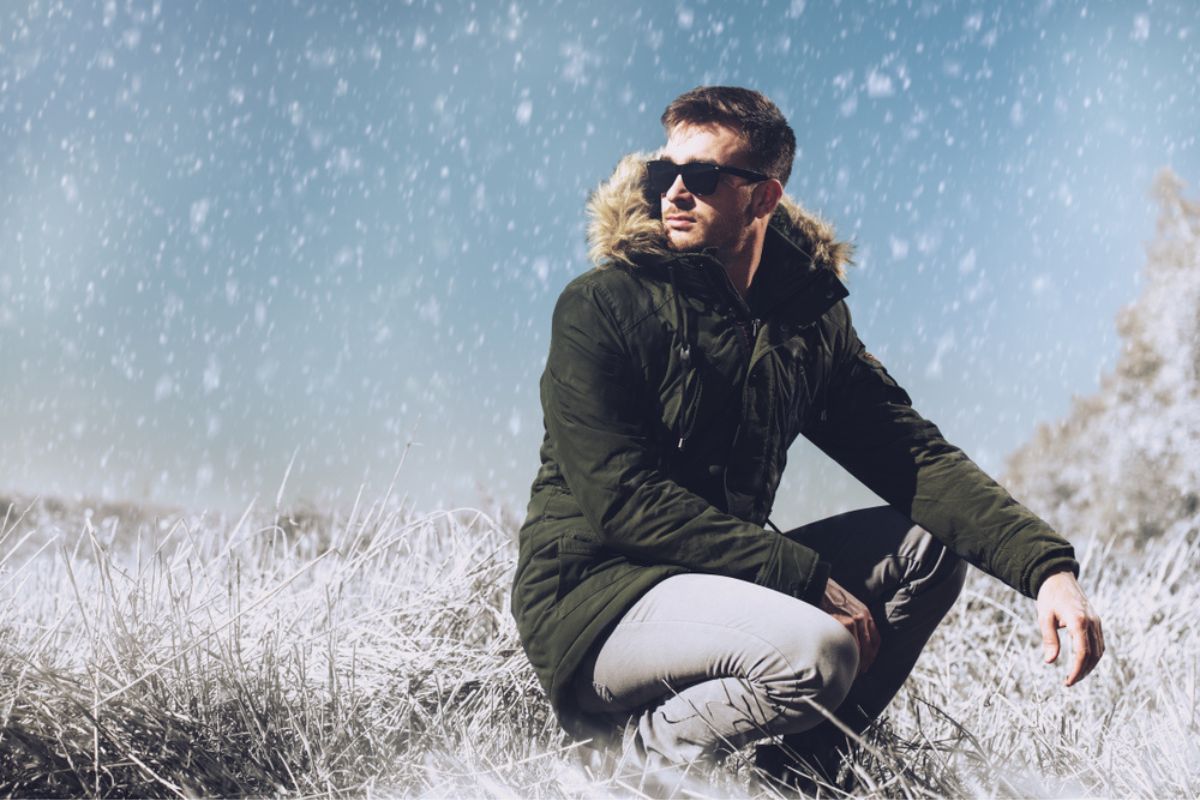A brutal handsome man wearing a parka and sunglasses in the countryside.